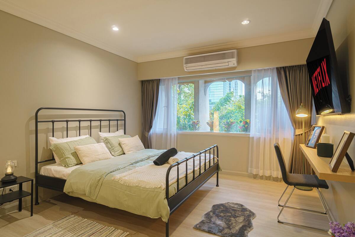 A bedroom with a bed, bedside table, air conditioning unit, window with curtains, and a desk with a chair.
