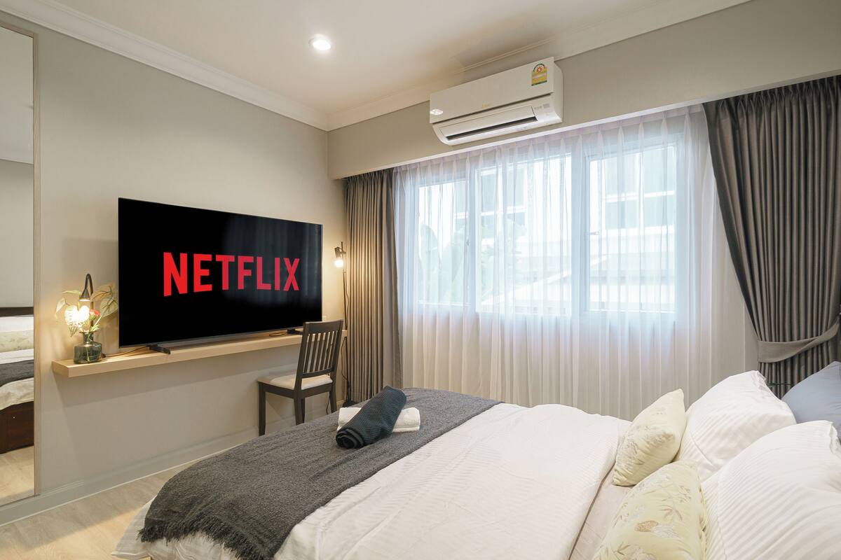 A hotel room with a large flat-screen TV mounted on the wall, a bed with white bedding, a bedside table with a lamp, and a window with curtains.