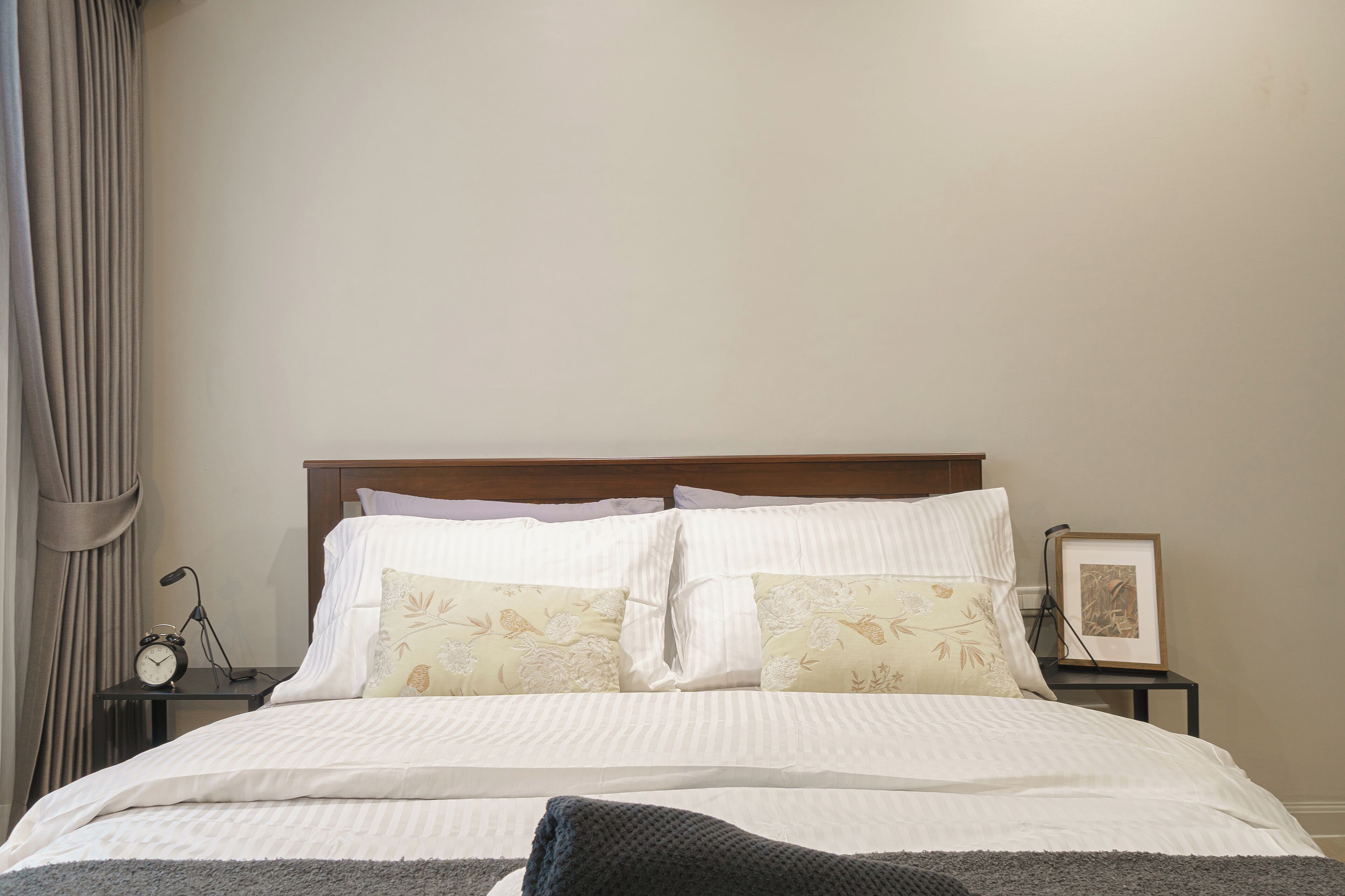 A neatly made bed with white linens and floral-patterned pillows, a wooden headboard, a bedside table with a clock, and a framed picture on the nightstand.