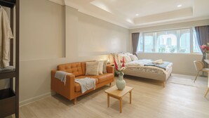 A modern living room with a sofa, a bed, a small table with a vase, and a view of buildings through large windows. - Casa Amor (Bangkok)
