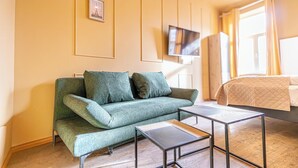 Een moderne kamer met een groene bank, een flatscreen televisie aan de muur, een bed met een beige deken en een raam met gordijnen.