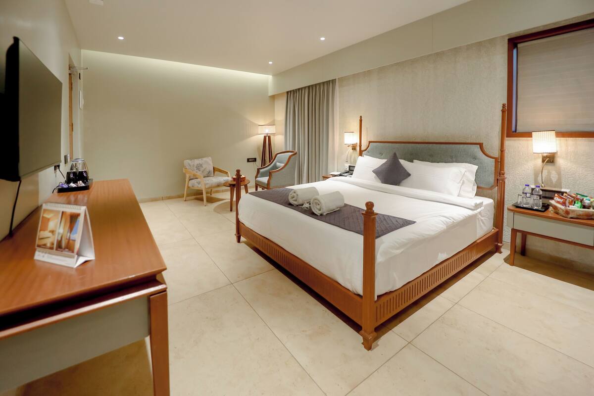 A hotel room with a wooden bed, a desk with a flat-screen TV, a chair, a nightstand with a lamp, and a window with curtains.