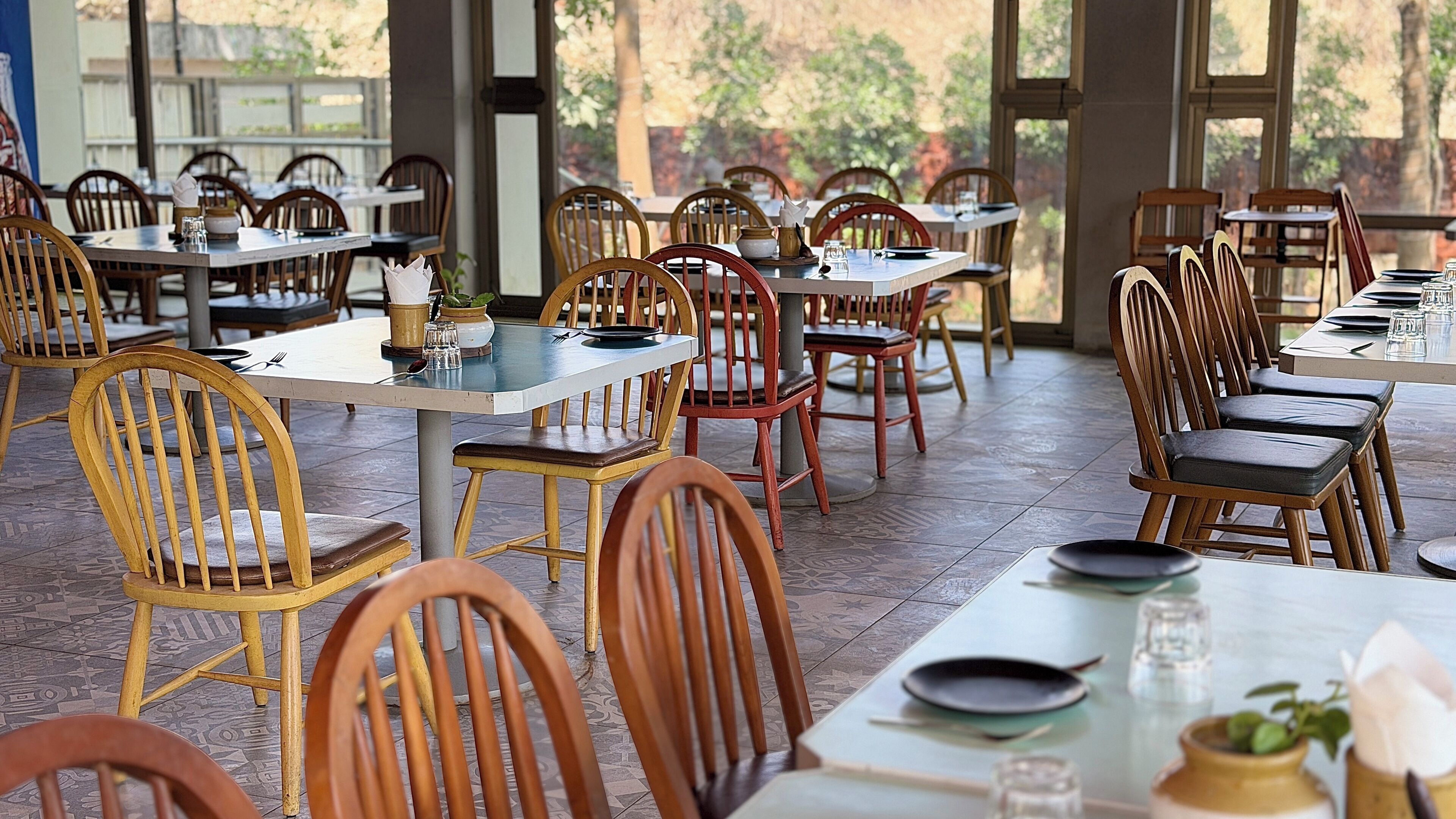 A restaurant with wooden chairs and tables, large windows, and a tiled floor.