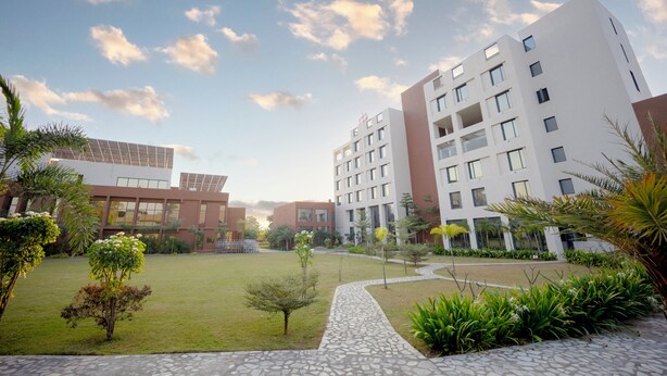 Un bâtiment moderne à plusieurs étages, une cour avec de la pelouse et des plantes, et un ciel dégagé avec des nuages.
