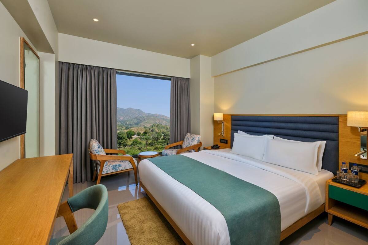A hotel room with a large bed, a desk, a chair, a TV, and a view of the mountains.