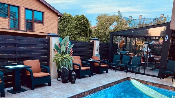 A swimming pool with lounge chairs and tables, surrounded by a wooden fence and a glass enclosure. - All Seasons Palanga (Palanga)