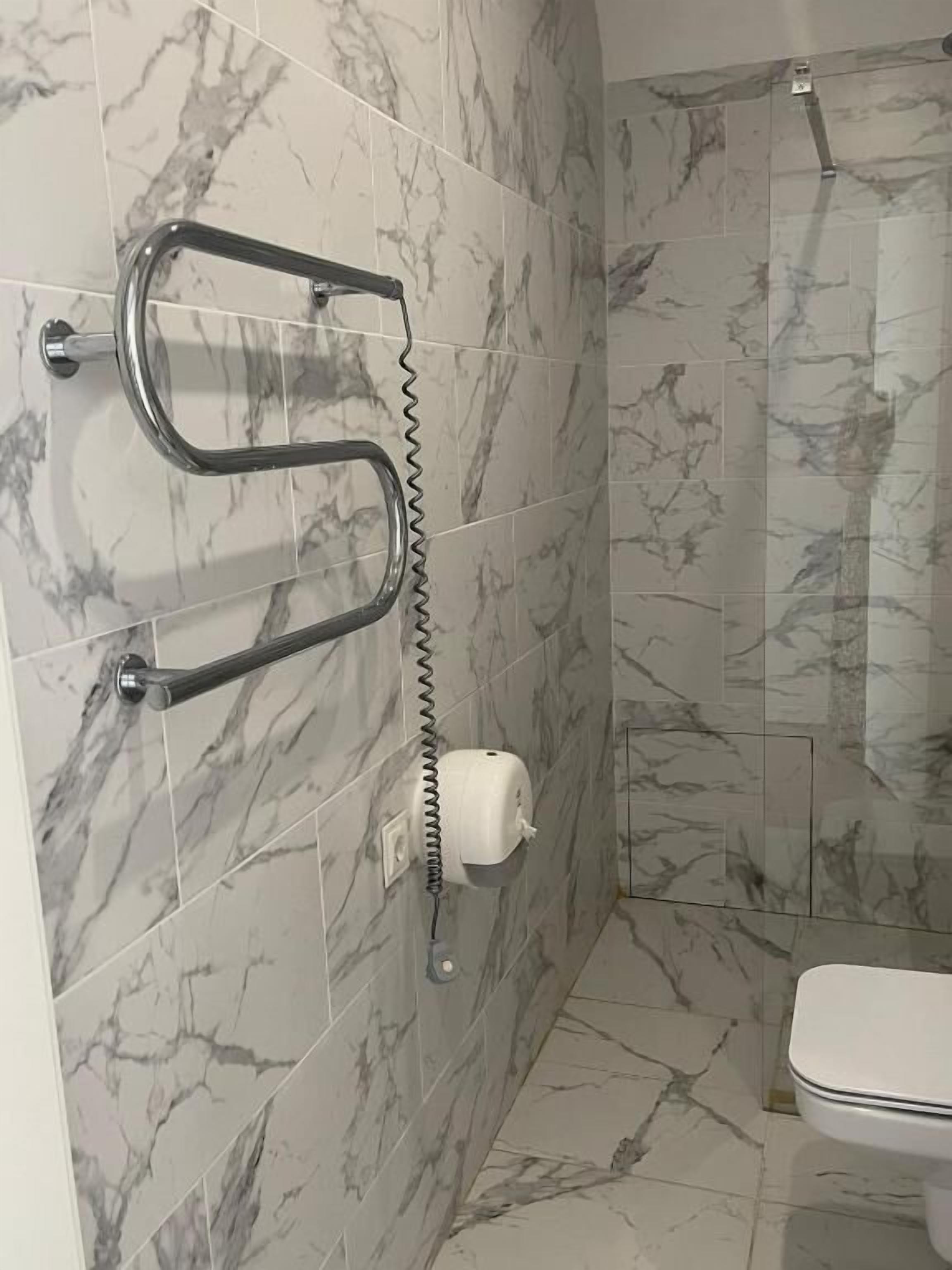 A bathroom with marble tiles, a toilet, a handrail, and a paper towel dispenser.