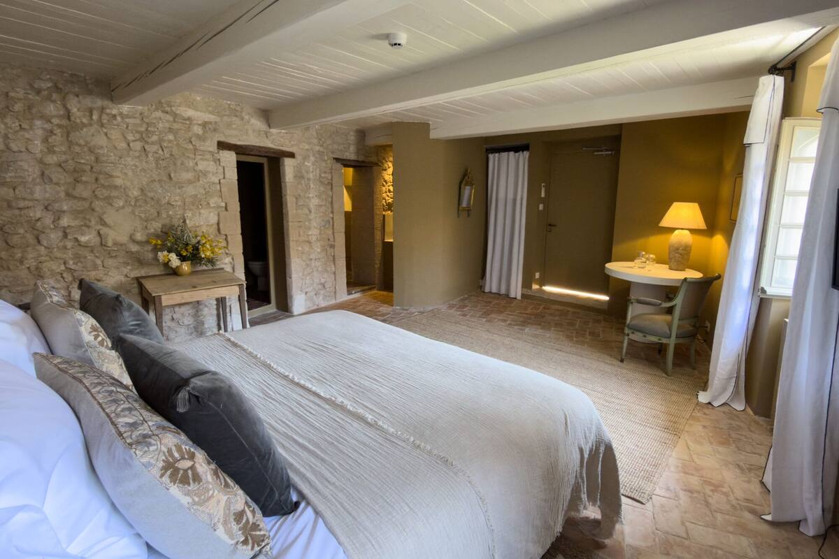 A bedroom with a large bed, a small table with a lamp, a bathroom with a shower, and a stone wall.