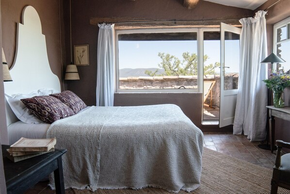 Un dormitorio con una cama grande, una mesita de noche, una lámpara, una ventana con cortinas y vistas al exterior.