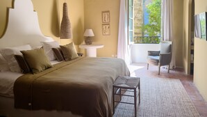 A bedroom with a large bed, a bedside table with a lamp, a chair, and a view of greenery outside. - Auberge du Presbytère (Saignon)