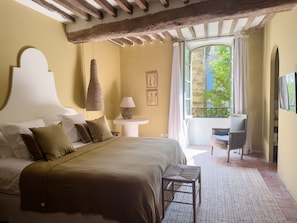 Quarto com uma cama grande, mesa de cabeceira com abajur, uma cadeira e vista para o verde lá fora.