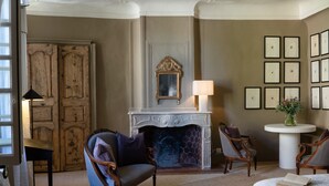 A room with a fireplace, framed artwork on the wall, and a round table with a vase. - Auberge du Presbytère (Saignon)
