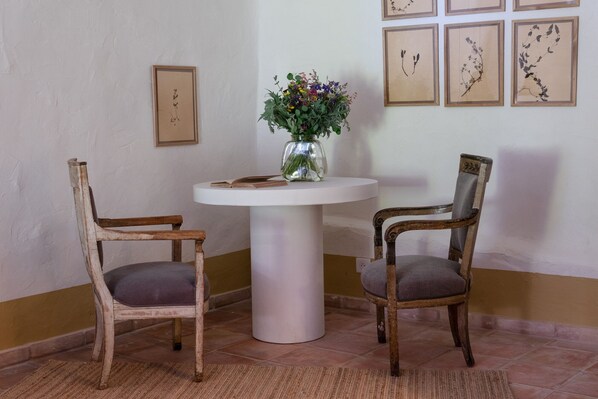Two wooden chairs with cushioned seats, a white pedestal table with a vase of flowers, and framed botanical prints on the wall.