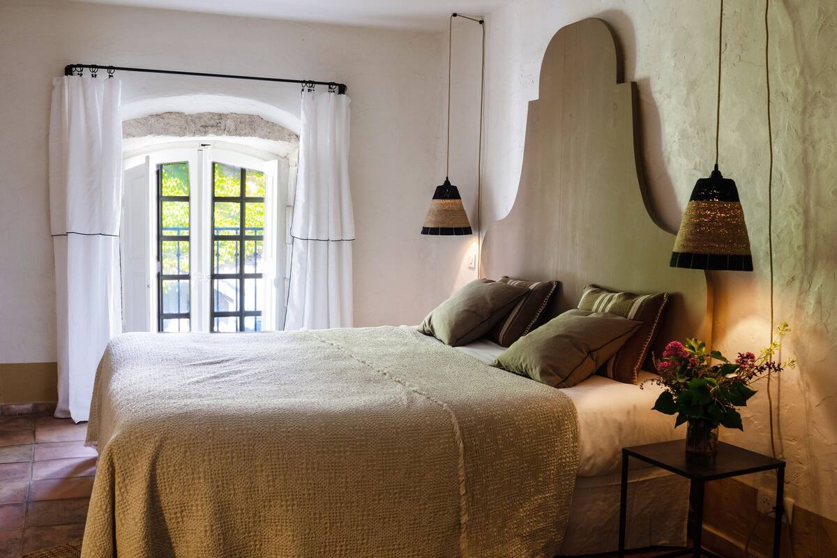 A bedroom with a bed, a nightstand, a window with curtains, and a wall-mounted lamp.
