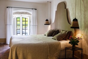 Un dormitorio con cama, mesita de noche, ventana con cortinas y lámpara de pared.