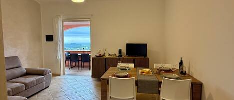 A living room with a sofa, a dining area with a table and chairs, a TV, and a view of the outdoors.
