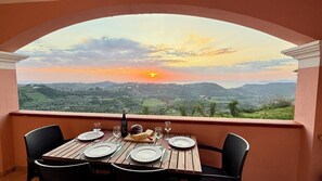 Une table dressée pour deux personnes, avec des assiettes, des verres et une bouteille, donnant sur un paysage magnifique illuminé par le coucher du soleil.