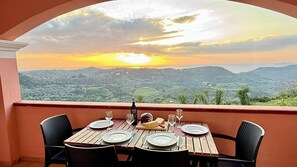 Une table dressée pour deux personnes, avec des assiettes, des verres, une bouteille et un morceau de fromage, donnant sur un paysage magnifique avec des montagnes et un coucher de soleil.