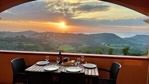 A table set for two with plates, glasses, and a bottle, overlooking a scenic landscape with hills and a sunset. - Sweet Holidays (Magomadas)