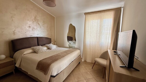 Une chambre à coucher comprenant un lit, un bureau, une chaise, un téléviseur et une fenêtre avec des rideaux.