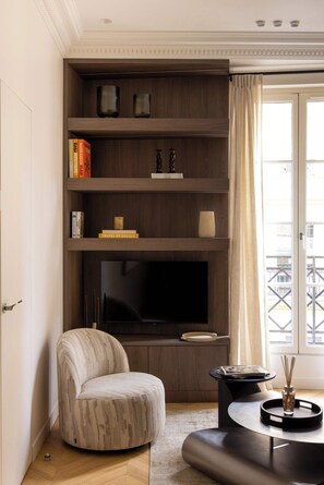 Een moderne woonkamer met een ingebouwde televisie, een comfortabele fauteuil, een klein bijzettafeltje en een boekenkast met boeken en decoratieve objecten.