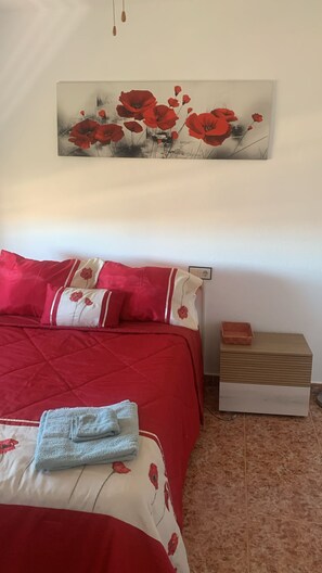 Une chambre à coucher avec une couvre-lit rouge, des coussins assortis et un tableau représentant des fleurs rouges au mur.