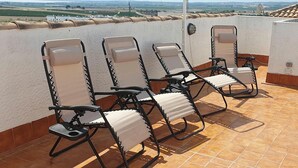 Quatre fauteuils inclinables avec repose-pieds, sur une terrasse carrelée, adossée à un mur blanc et offrant une vue sur la campagne.