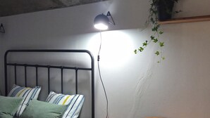 A small, dimly lit room with a metal bed frame, a bedside table, and a small stool. A single lamp provides light, and a potted plant hangs on the wall. - Adega de Pedra, charming, renovated old winery in the north of Madeira (Boaventura)