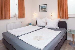 A double bed with white linens, a bedside table with a cup and a book, and a window with curtains. - Elsa - Wenningstedt-Braderup (Sylt) (Wenningstedt-Braderup (Sylt))