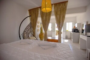 A bedroom with a bed, a chair, curtains, a hanging lamp, and a view of a kitchen area. - Rhapsody Traditional Apartments (Santorini)