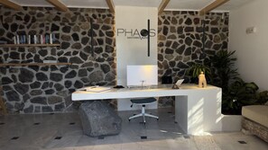 A reception desk with a stone wall backdrop, a mounted TV, and a small statue. - Rhapsody Traditional Apartments (Santorini)