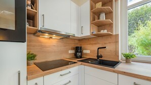 A modern kitchen with white cabinets, a black sink, and a built-in stove. There are wooden shelves above the counter and a window with a view of greenery. - Villa Sabinium 01: Close to the beach in Heringsdorf with your own beach chair (Heringsdorf (Seebad))