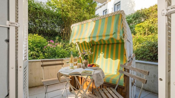 An outdoor balcony with a table set for two, a striped awning, and a view of greenery and a building in the background. - Villa Sabinium 01: Close to the beach in Heringsdorf with your own beach chair (Heringsdorf (Seebad))