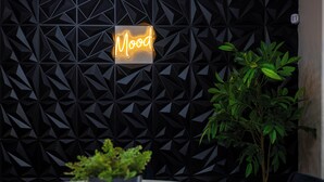 Ein modernes Interieur mit einem geometrischen Wanddesign, einem Neon-Schild mit der Aufschrift "Mood", einem runden Tisch mit einer Topfpflanze und einer hohen, grünen Pflanze neben der Wand.