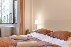 Una cama bien tendida con una colcha blanca y rosa, una lámpara en la mesita de noche y vista a un edificio por la ventana.