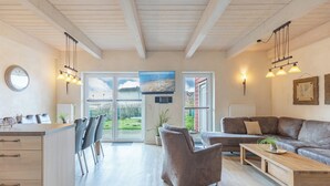 En rummelig opholdsstue med træloft, stor sofa, sofabord og et køkkenområde med bord og stole.