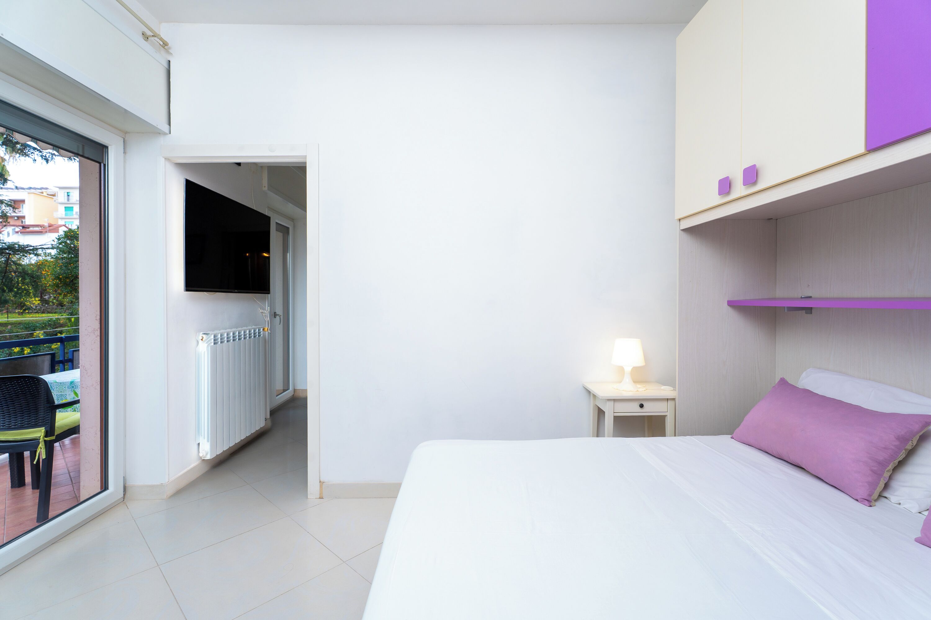 Un dormitorio con cama, mesita de noche, televisor y ventana con vistas a un balcón.