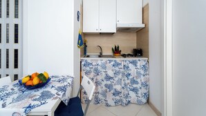 Cocina pequeña con armarios blancos, un fregadero y una estufa. Se ve una mesa con mantel azul y un cuenco de frutas.