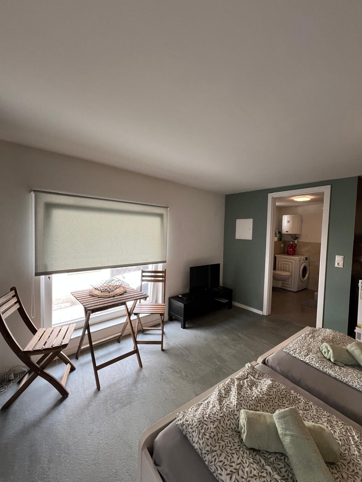 A compact room with a bed, a small table with chairs, a TV, and a window with blinds.