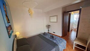 Quarto com uma cama, mesa de cabeceira, abajur, ventilador na parede e porta para outro cômodo.