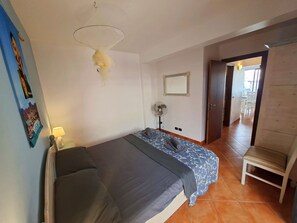 Ein Schlafzimmer mit einem Bett, einem Nachttisch, einer Lampe, einem Wandventilator und einer Tür zu einem anderen Raum.