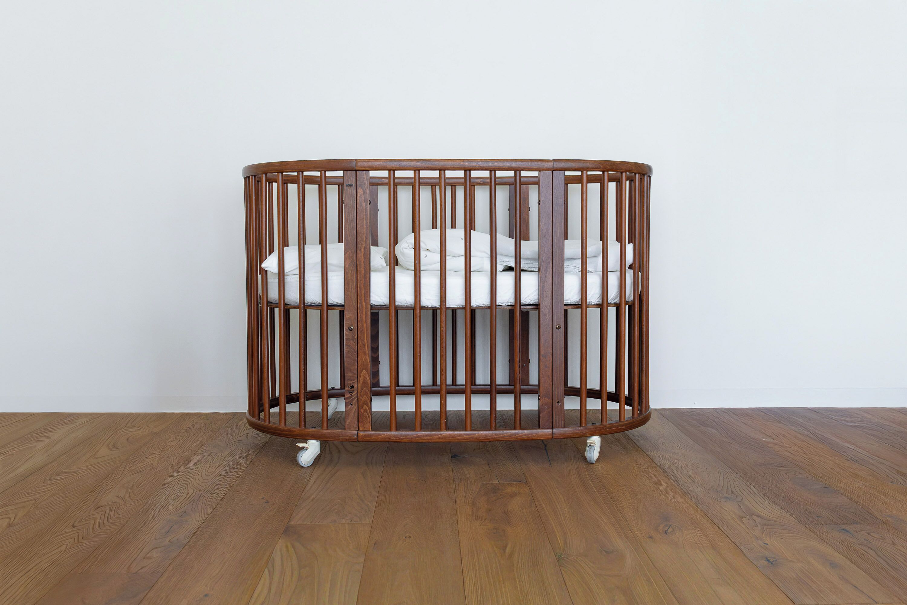 A wooden crib with a curved design, positioned on a wooden floor against a plain wall.