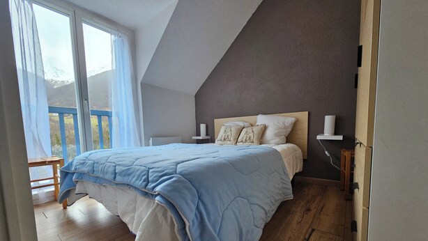 Een slaapkamer met een bed, houten vloer, een raam met gordijnen en uitzicht op het landschap.