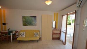 Een woonkamer met een bank, een tafel en stoelen. Aan de muur hangt een schilderij en aan het plafond bevindt zich een airconditioner.