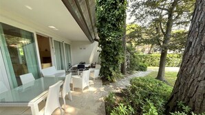 Zona pranzo all'aperto con tavolo in vetro e sedie bianche, circondata da vegetazione e un albero.