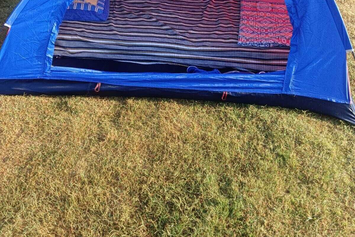 Uma tenda azul com um tapete estampado no interior, instalada numa área gramada com um muro de tijolos e uma estátua de urso ao fundo.
