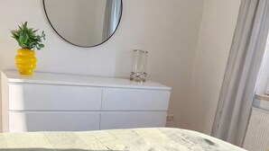 A bed with a grey and white patterned bedspread, a dark grey pillow, and a yellow accent pillow. A white dresser with a round mirror above it and a small potted plant on top is visible. - Luxury apartment - Charming 2 room apartment (Augsburg)