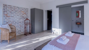 Una habitación de hotel moderna con pared de ladrillo, una cama grande con toallas, una silla de mimbre y una mesita de madera.