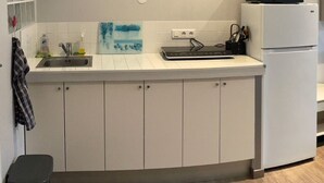 Eine Küche mit weißen Schränken, einer Edelstahl-Spüle, einer Mikrowelle auf einem weißen Kühlschrank und einer schwarzen Mülltonne.
