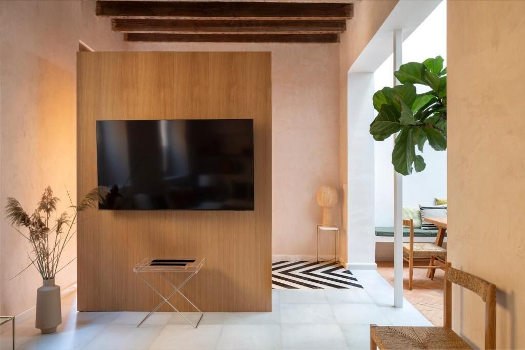 Ein modernes Wohnzimmer mit einem großen Flachbildfernseher, der an einer Holzwand angebracht ist, einem kleinen Beistelltisch mit Lampe, einer üppigen Topfpflanze und einem schwarz-weißen Teppich mit Muster.